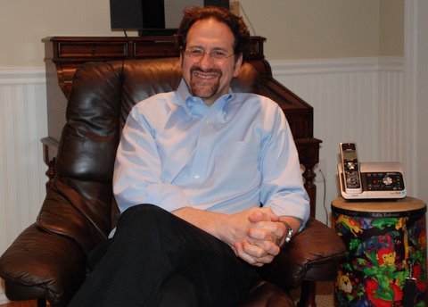 The image features a man sitting in a chair, smiling at the camera. He is wearing a blue shirt and has his arms crossed on his lap. The background includes a couch and what appears to be a wooden table with a remote control on it.