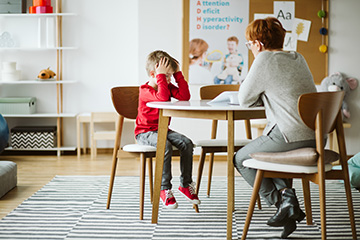 An adult and a child sitting at a table in a modern living room, with the adult holding their head in their hands.