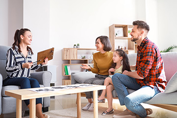 A family in a modern living room, with two adults and a child seated on a couch, engaged in a conversation with another adult standing nearby, while one person is holding a book.