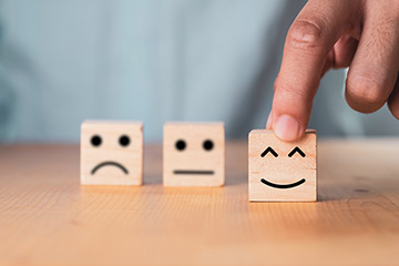 The image shows a person s hand placing a smiley face block into the first position of four blocks on a light-colored surface, with three other blocks in front of it, each featuring a different facial expression.