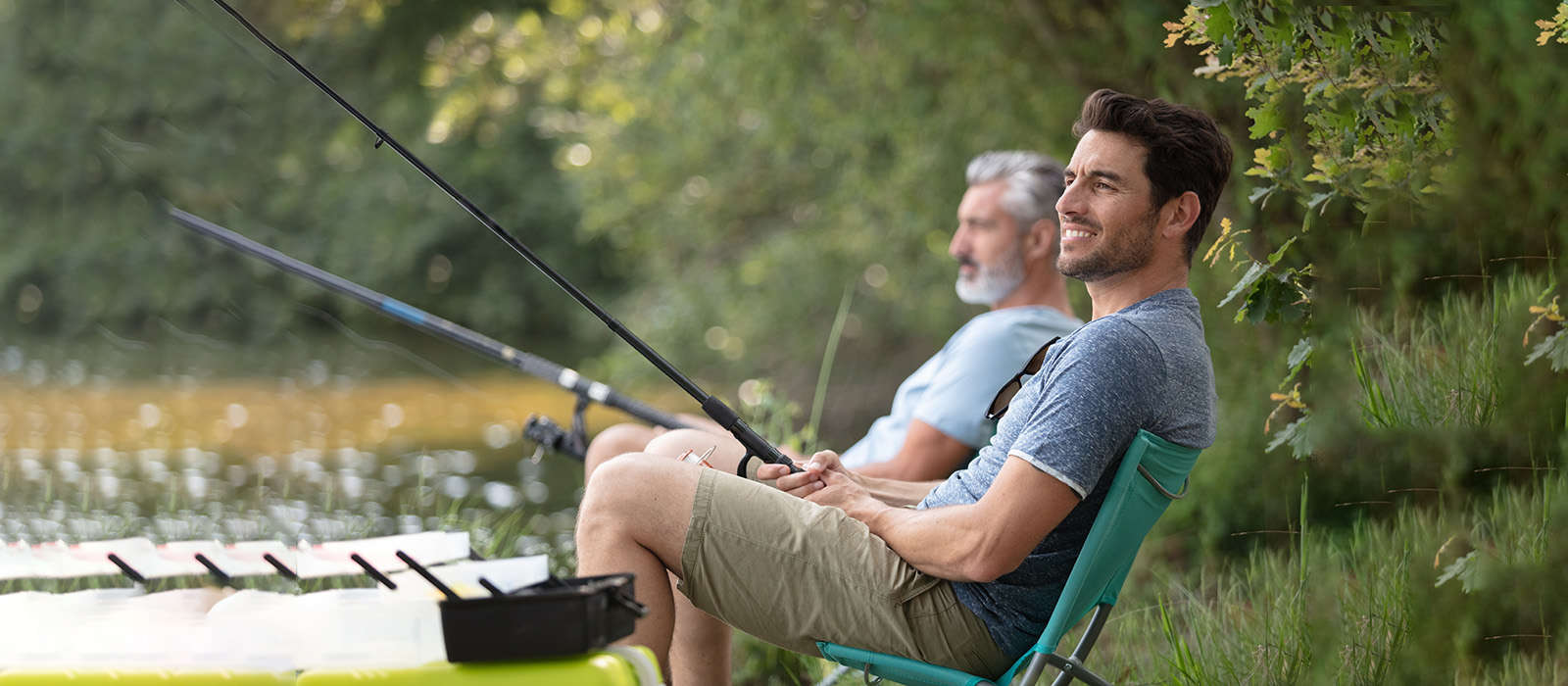 Two men sitting in folding chairs on a riverbank, one holding a fishing rod, with a body of water and lush greenery in the background.